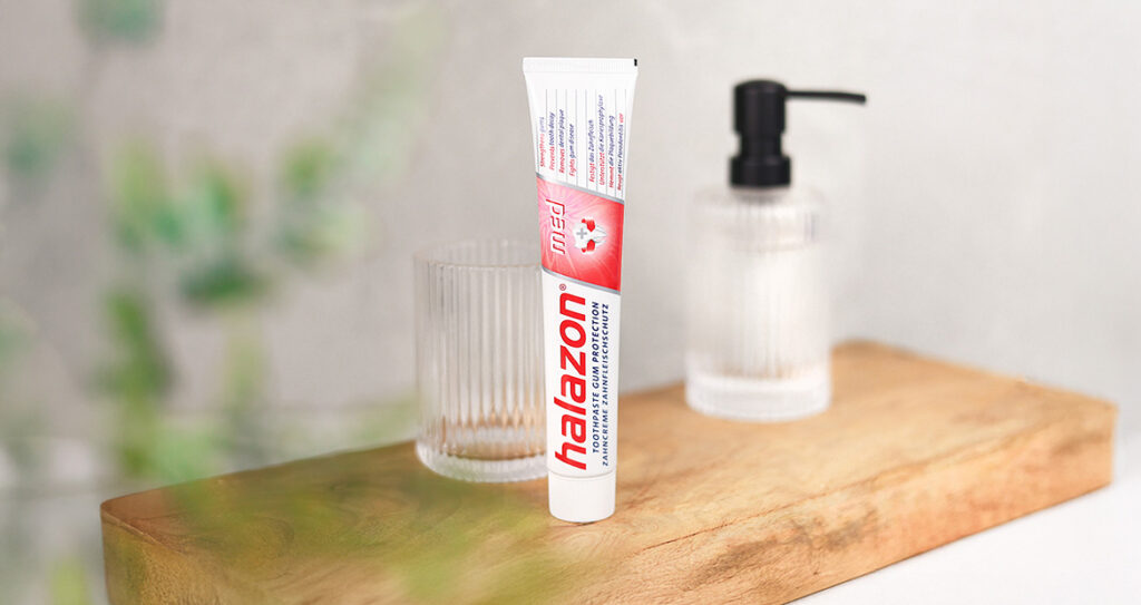 Eine Tube Halazon Zahnpasta steht auf einem Holzbrett neben einem Seifenspender und einem Glas, ideal für Mundhygiene.