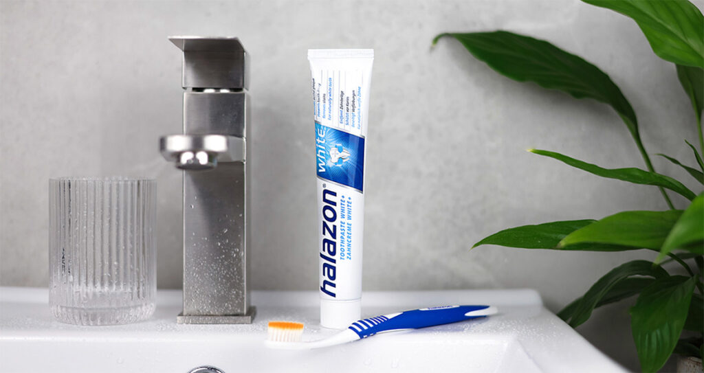 Eine Tube Zahnpasta mit der Aufschrift "Halazon White" steht neben einem Zahnputzbecher und einer Zahnbürste auf einem Waschbecken.