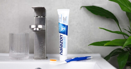 Eine Tube Zahnpasta mit der Aufschrift "Halazon White" steht neben einem Zahnputzbecher und einer Zahnbürste auf einem Waschbecken.