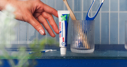 Eine Hand greift nach einer Tube Zahncremekonzentrat von One Drop Only auf einem Badezimmerregal für optimale Mundhygiene.